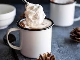 White mug topped with whipped cream and cocoa on a dark table, with pinecones nearby