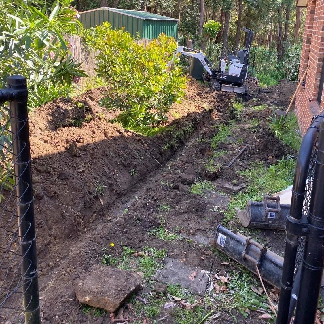 A trench dug in a backyard garden next to a black fence with a small excavator parked near a green shed.