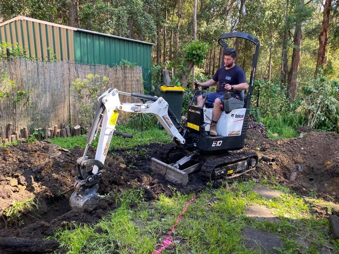 A person operating a small white and black excavator to dig a trench in a yard near a green metal shed.