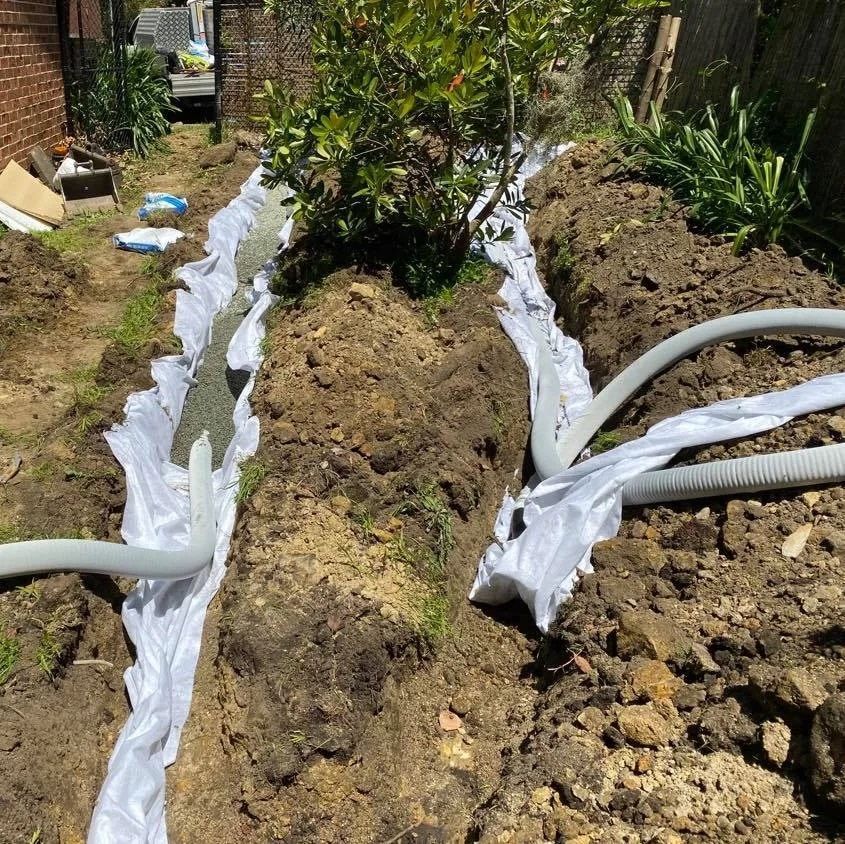 A backyard trench lined with white landscape fabric, containing gravel and a flexible black drainage pipe.