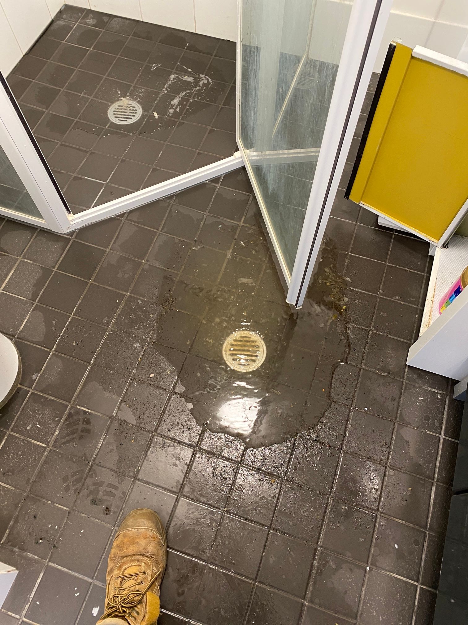 A wet bathroom floor with a puddle surrounding a floor drain near a shower enclosure, viewed from a high angle.