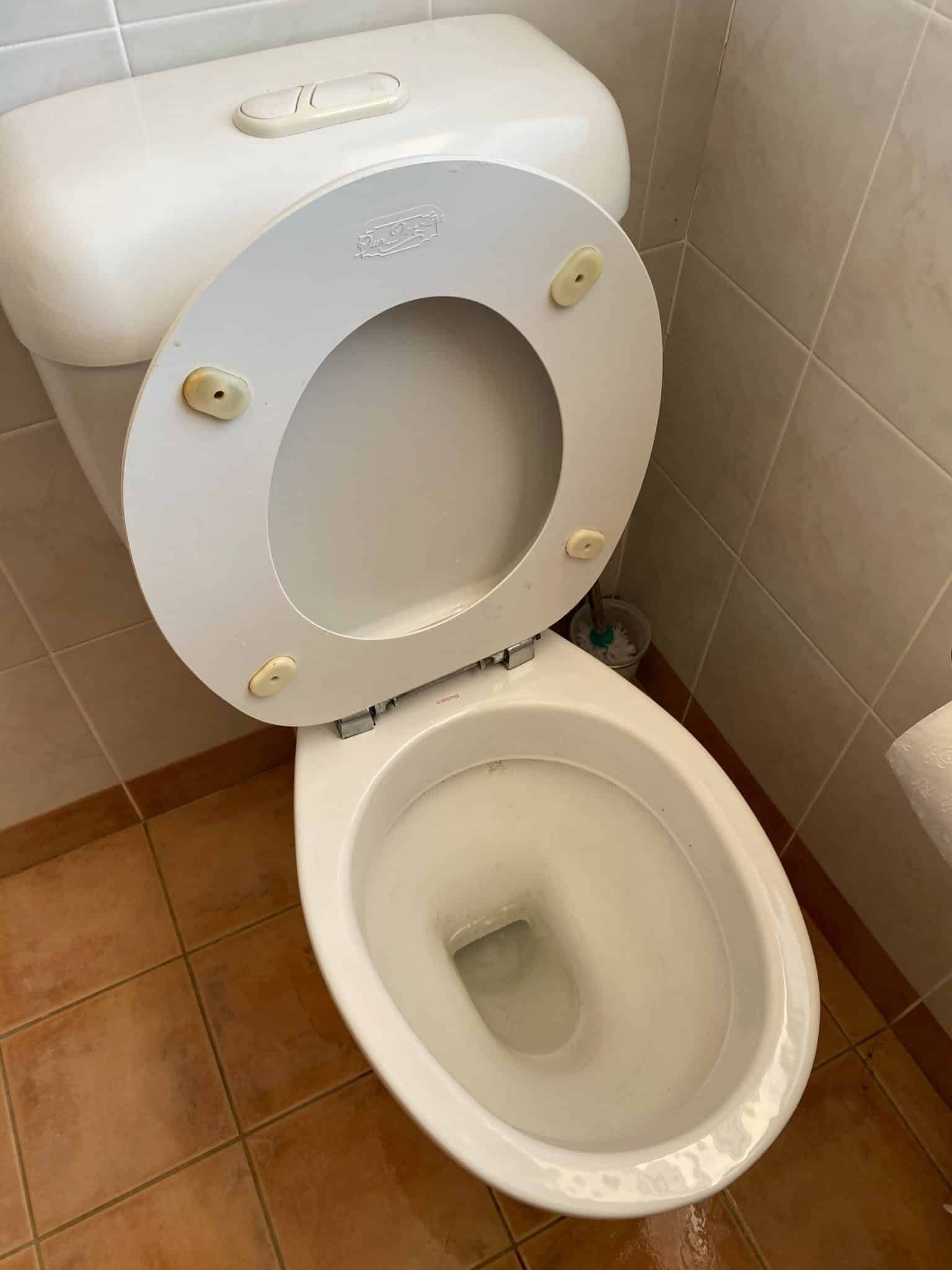 A white toilet with the seat lid raised, positioned in a bathroom with beige tiled walls and floor.