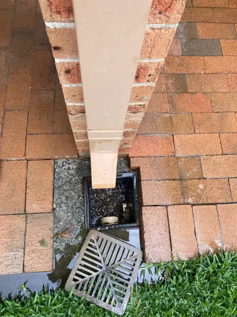 A downspout positioned above an open, square storm drain set in a brick patio, with the metal grate lying beside it.