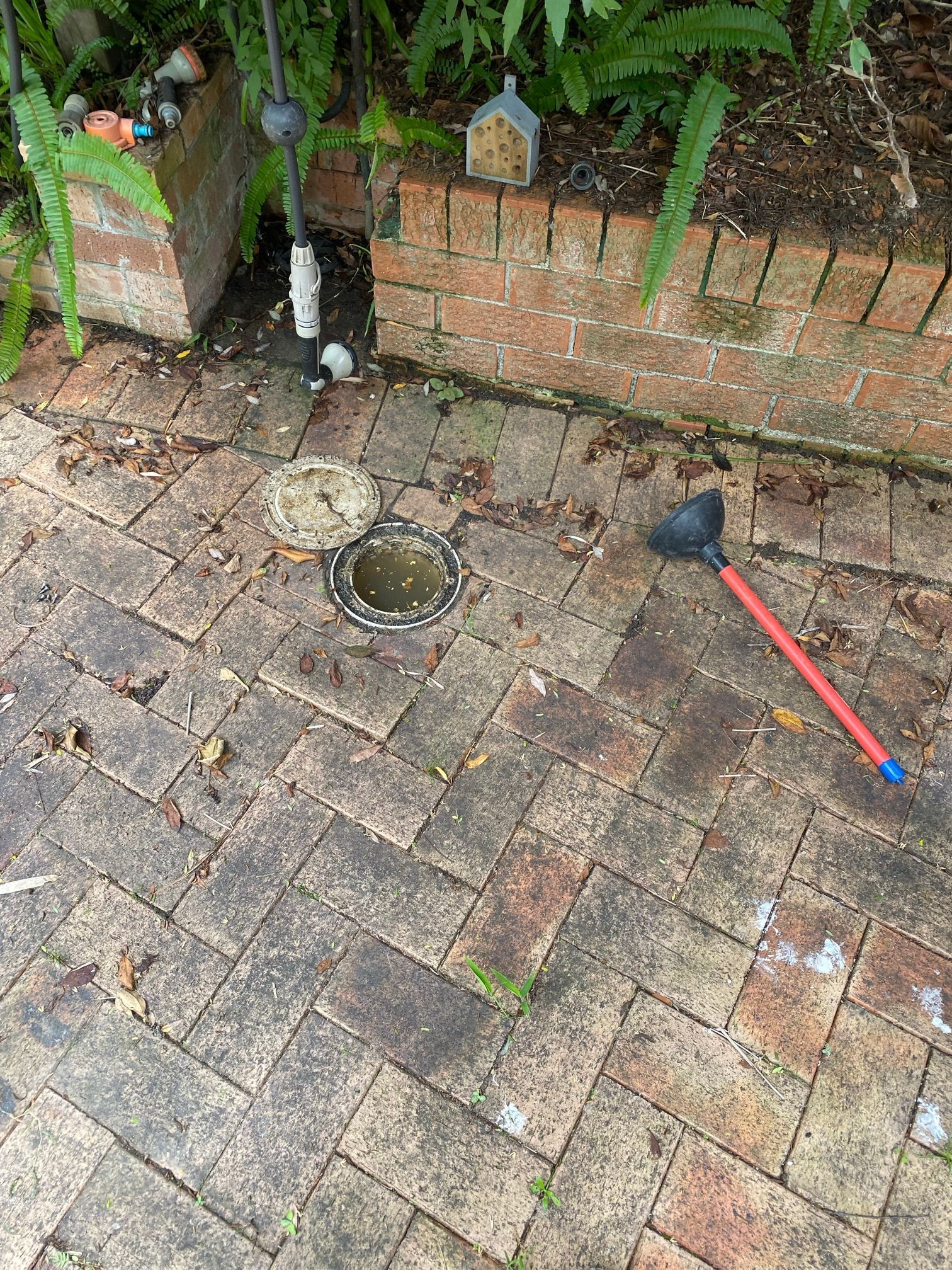 A drainage inspection point with its lid open, set into a red brick paved patio near a brick garden wall with a plunger.