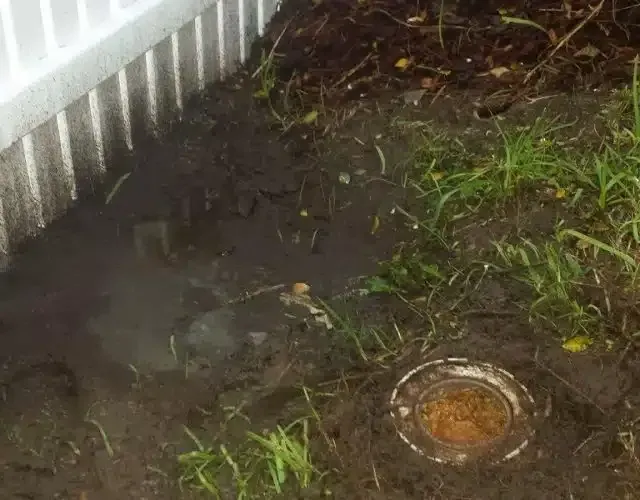 A muddy patch of ground beside a white fence with a circular drainage pipe or access cap visible in the grass.