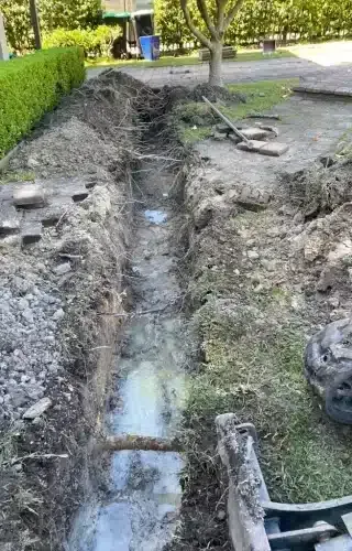 A long, muddy trench dug into a residential lawn, with water pooling at the bottom and excavation equipment in the corner.
