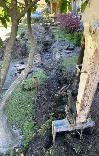 An excavator bucket rests in a freshly dug trench in a residential front yard with lawn and landscaping nearby.