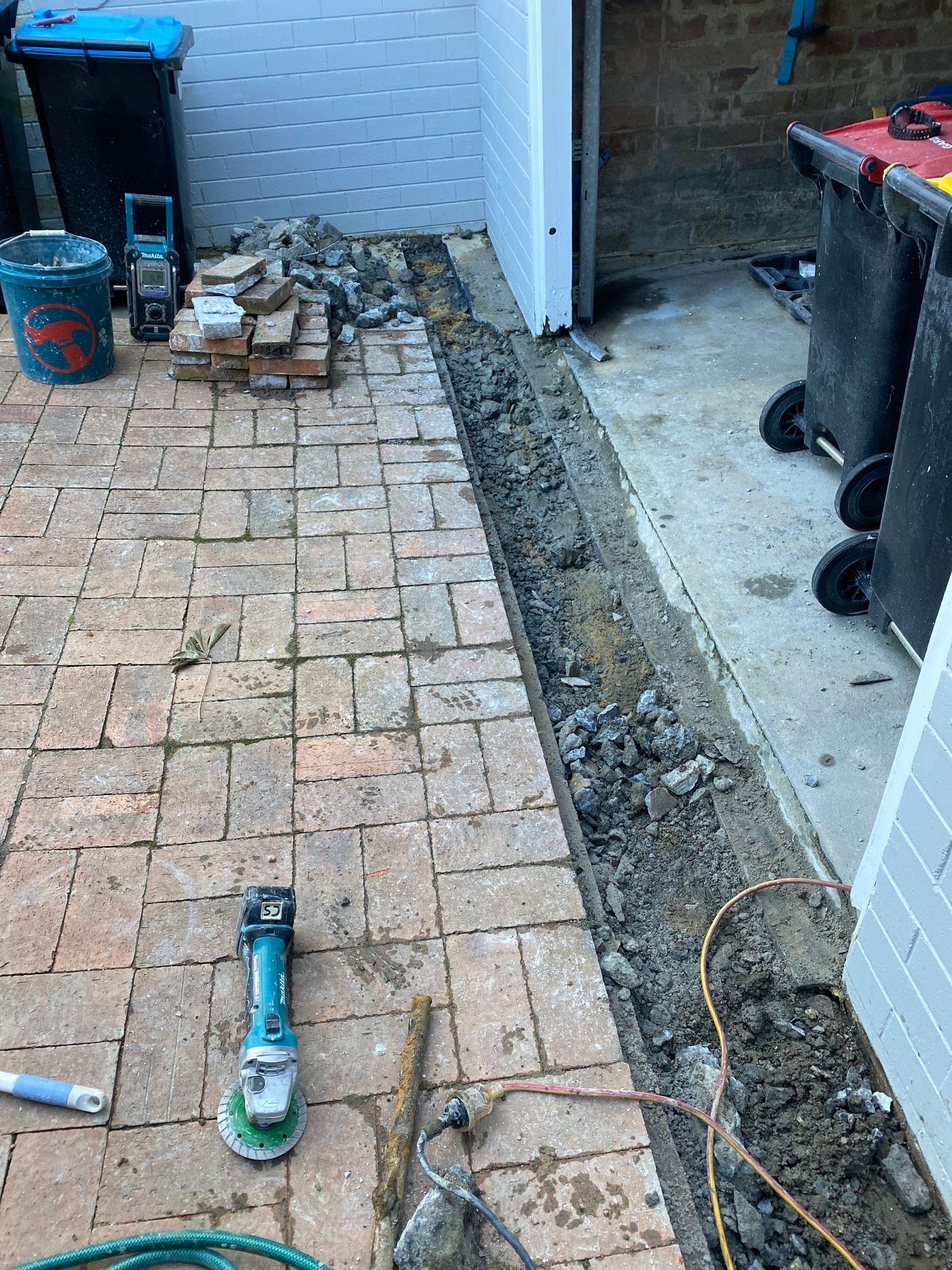 A narrow trench dug through brick pavers in a driveway, with a power tool, bricks, and trash bins nearby.
