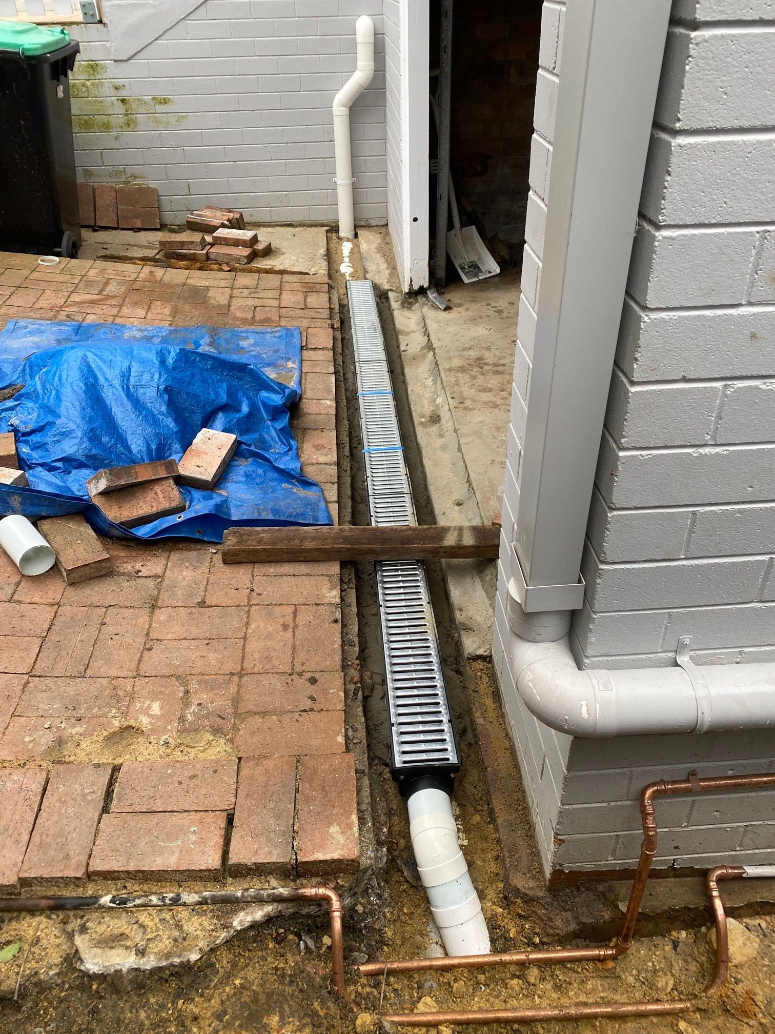 A trench drain with a metal grate installed along a gray brick wall, with exposed copper pipes and brick pavers nearby.