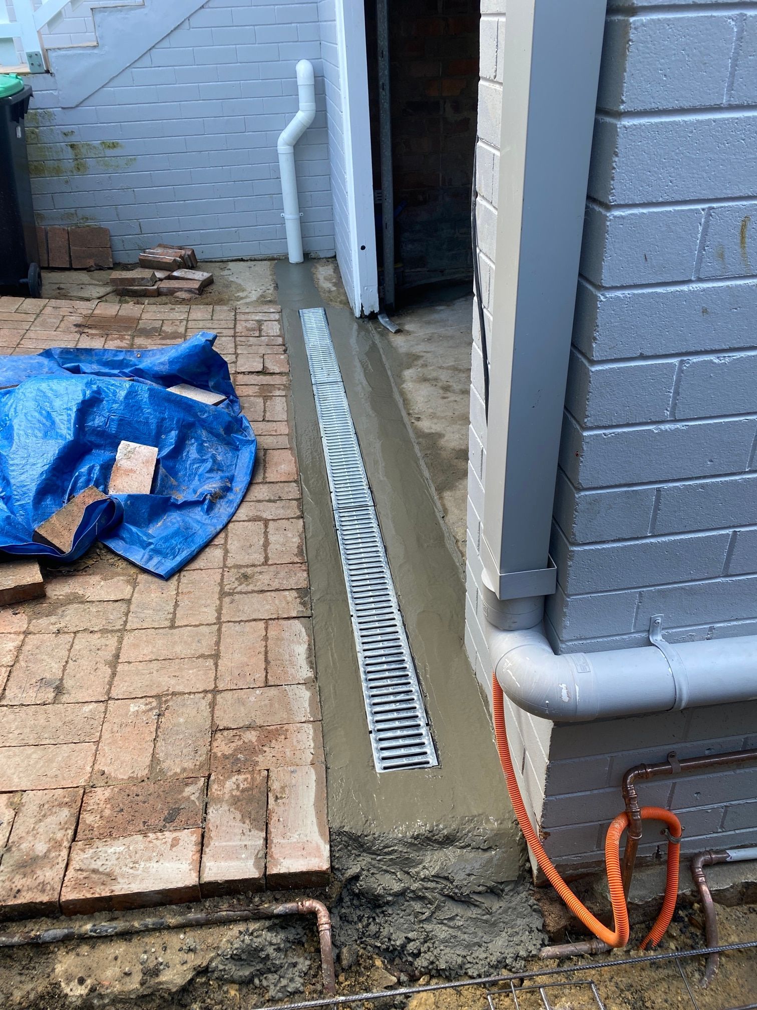 A newly installed grey trench drain set in fresh concrete alongside a brick wall and a paved patio area.
