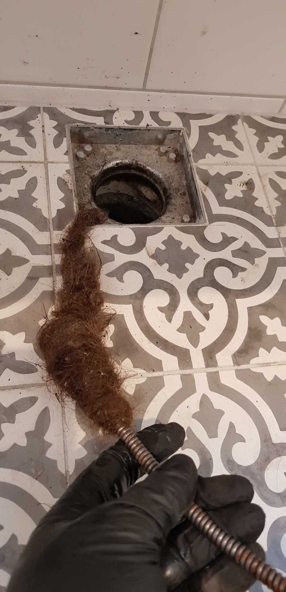 A gloved hand holding a drain cleaning tool coated with a thick, matted clump of hair removed from a tiled shower floor.