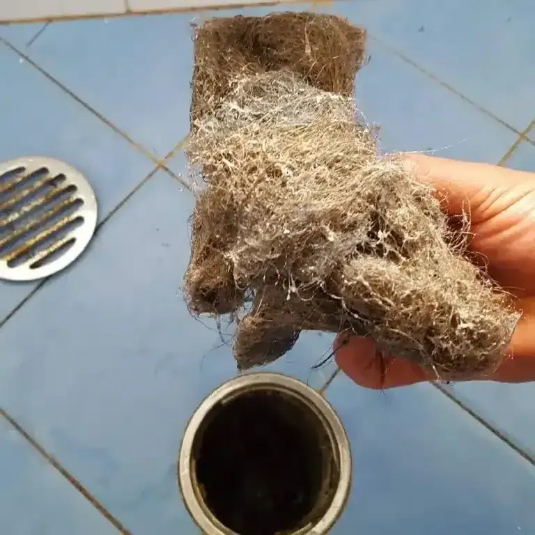 A hand holds a large clump of wet hair and debris removed from a bathroom drain, with the open drain visible below.