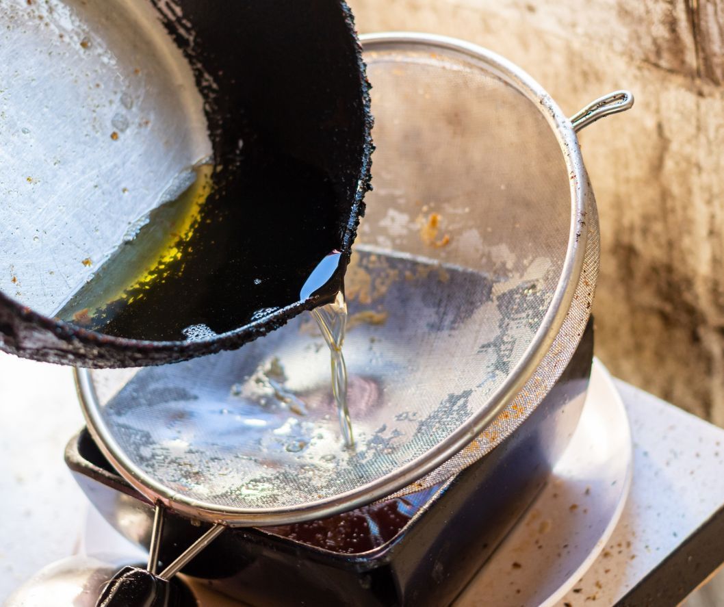 Cooking oil being poured from a dark pan through a metal mesh strainer into a container.