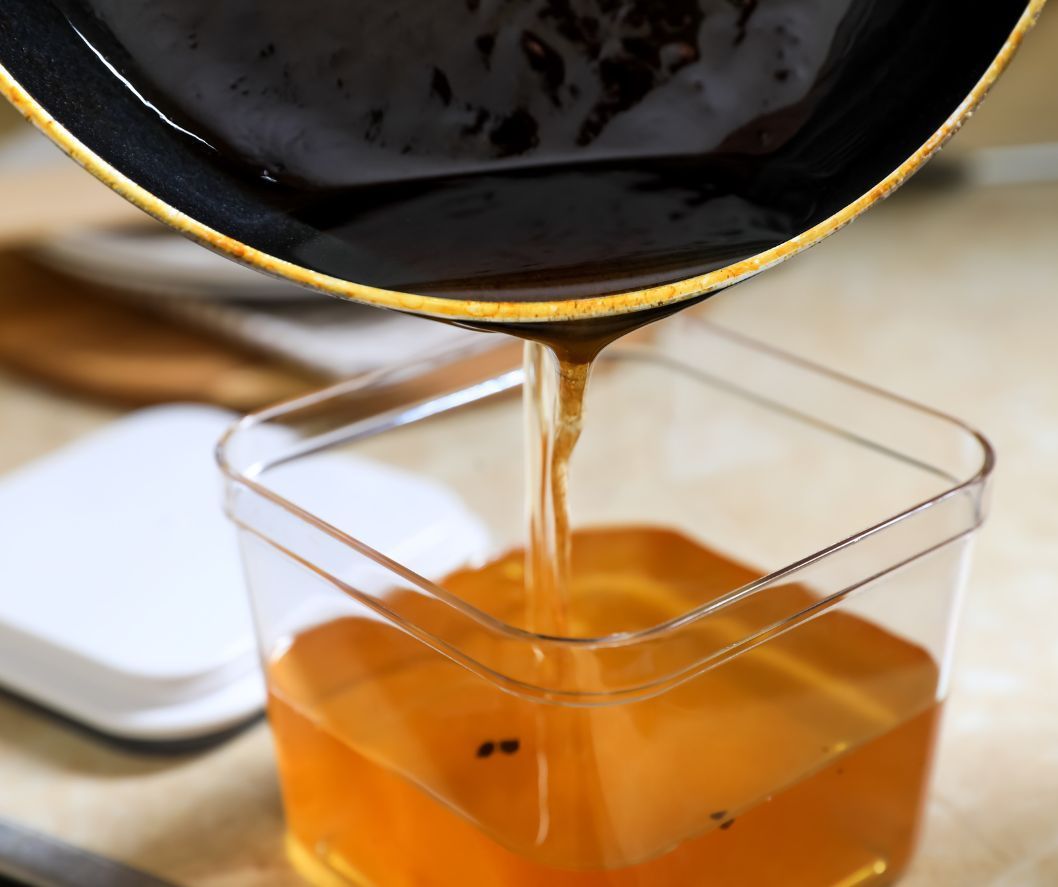 A dark liquid being poured from a pan into a clear, square container filled with a translucent, golden-amber liquid.