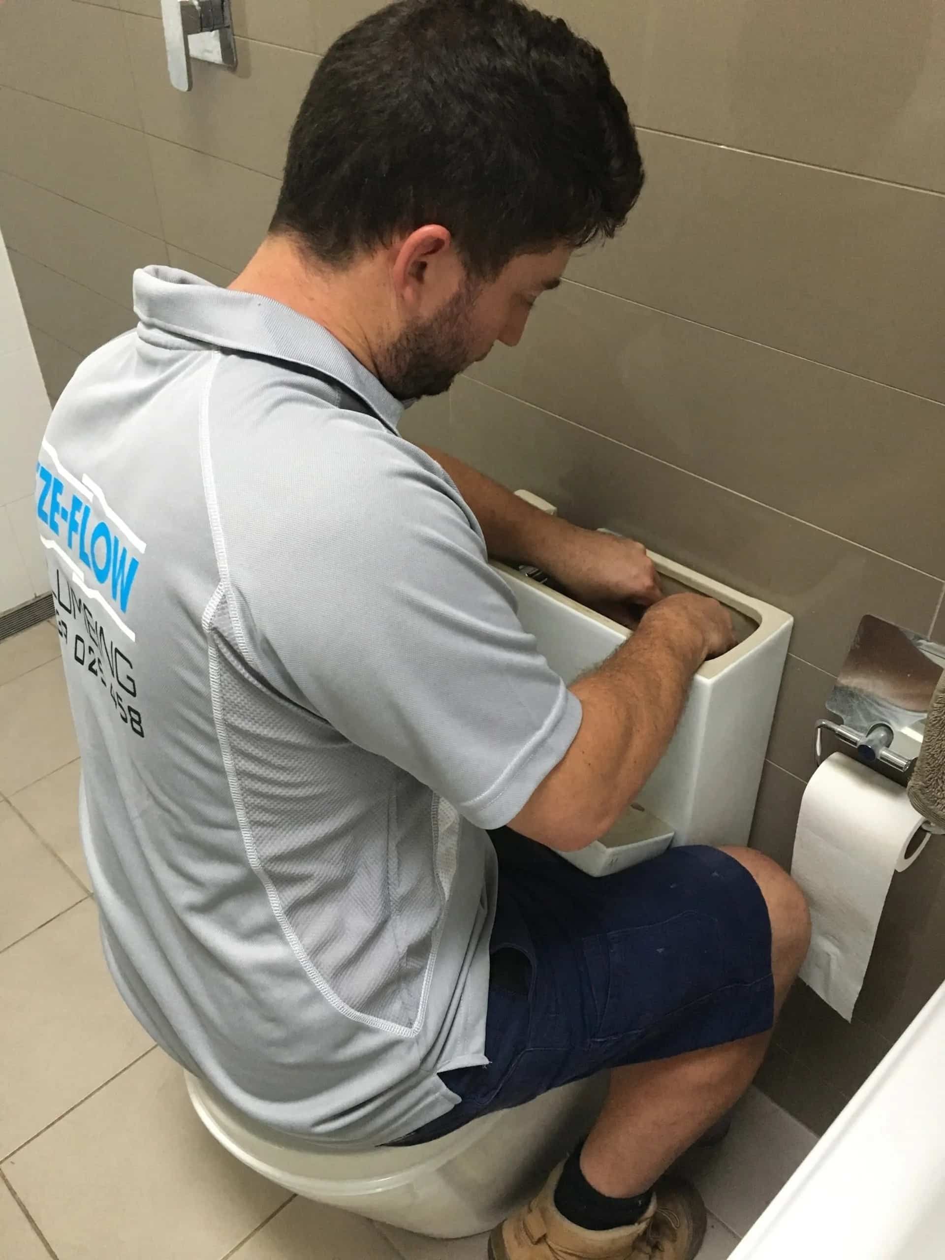A person in a gray service uniform repairs the internal mechanism inside an open toilet tank in a tiled bathroom.