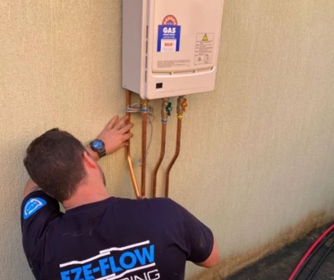 A technician in a blue uniform installs copper plumbing pipes onto a wall-mounted gas water heater outdoors.