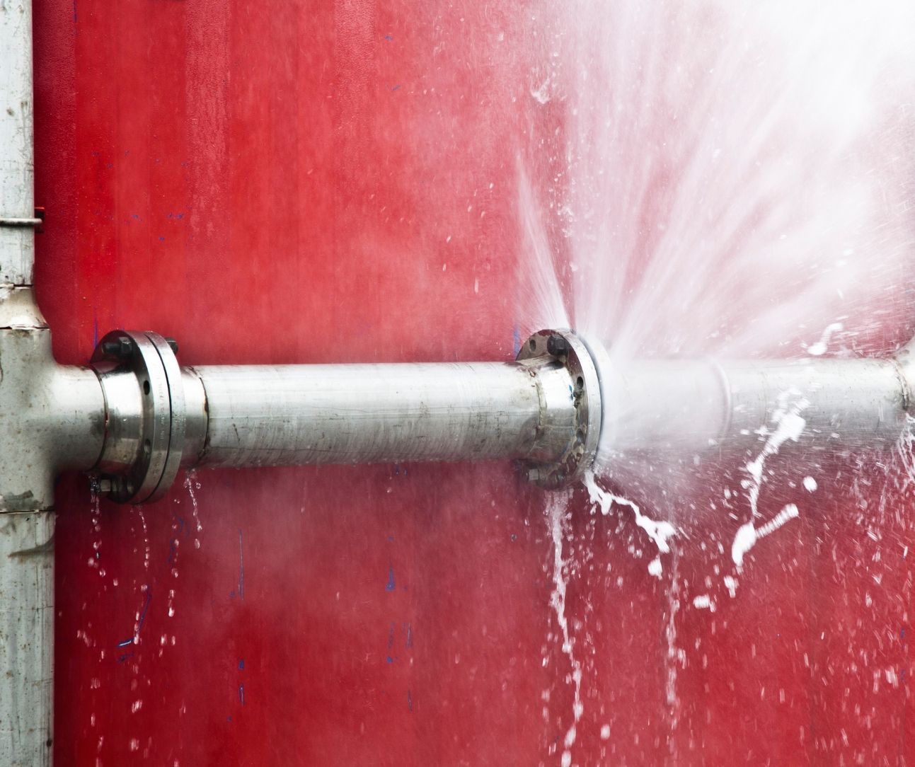 A high-pressure water spray leaks from the flanged connection of a silver metal pipe against a bright red background.