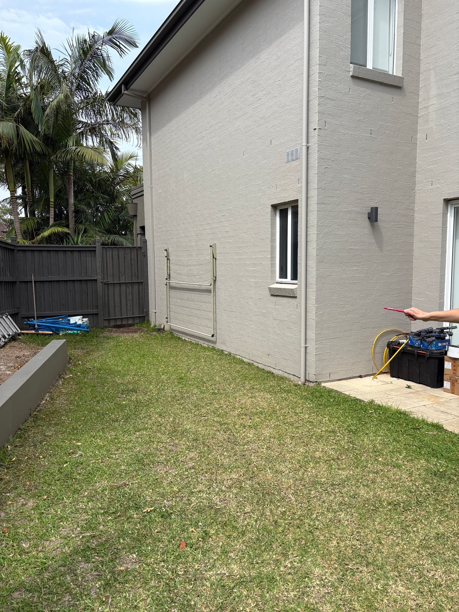 A light-colored stucco side wall of a house with a window, facing a grassy lawn and a wooden fence.