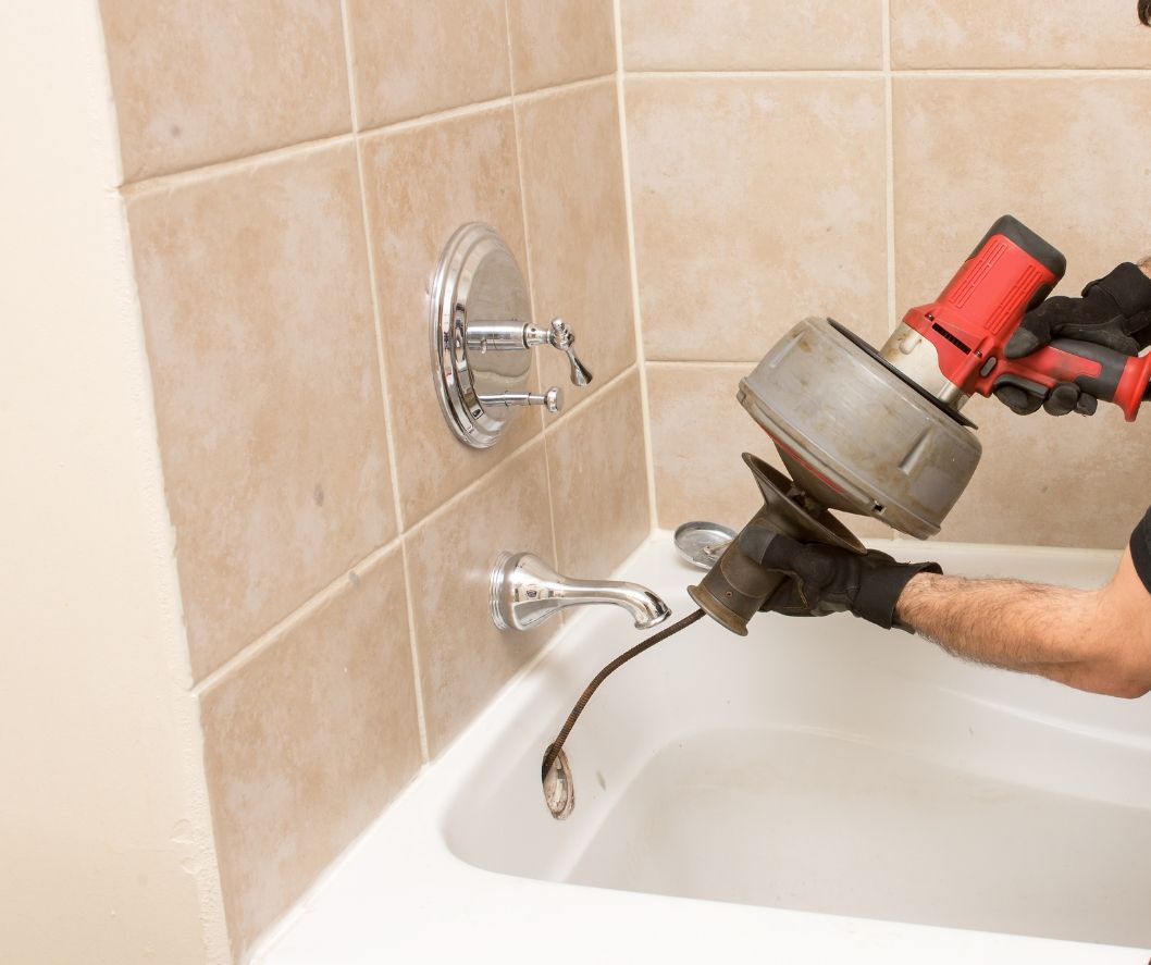 A plumber in black gloves uses a power drain auger to clear a clog in a white bathtub drain.