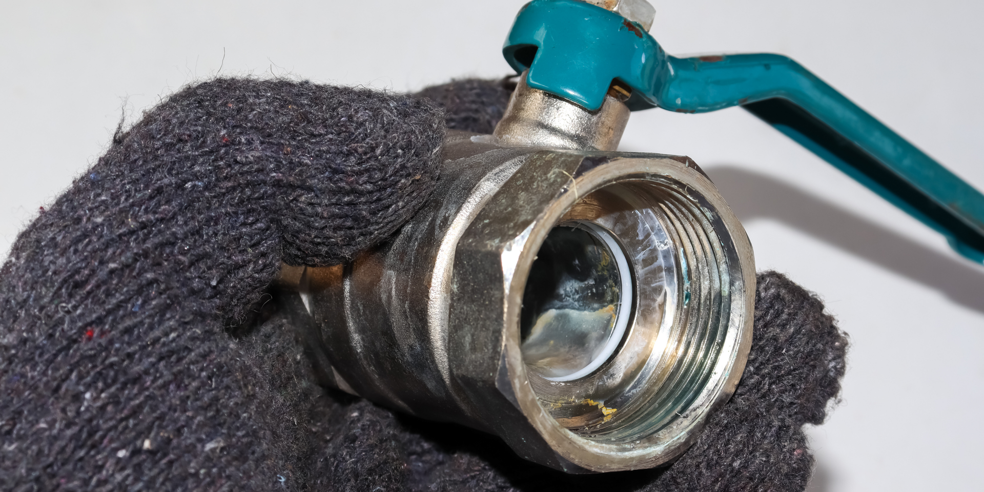 Gloved hand holding a silver ball valve with a teal lever handle, close-up on threaded opening Gloved hand holding a silver ball valve with a teal lever handle, close-up on threaded opening