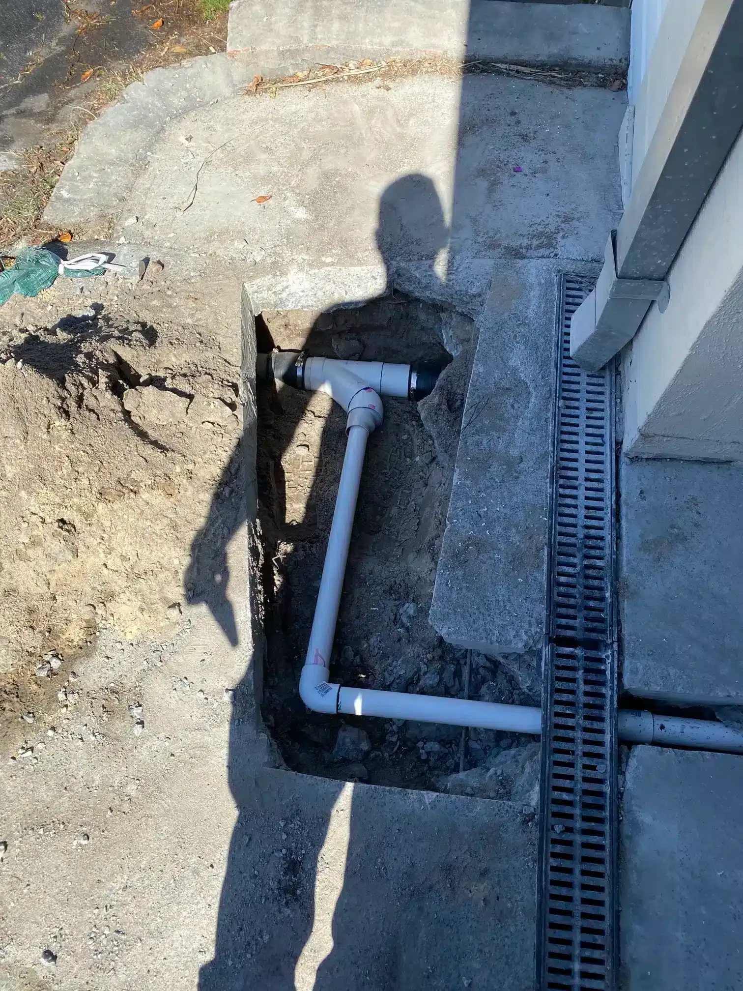 A trench in concrete reveals a white PVC pipe assembly connected to a drainage grate near a building foundation.