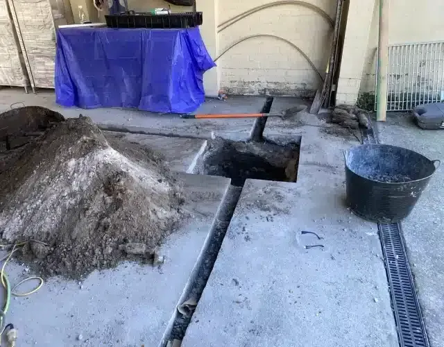 A large pile of dirt sits next to a rectangular hole in a concrete floor, with a shovel and a black bucket nearby.
