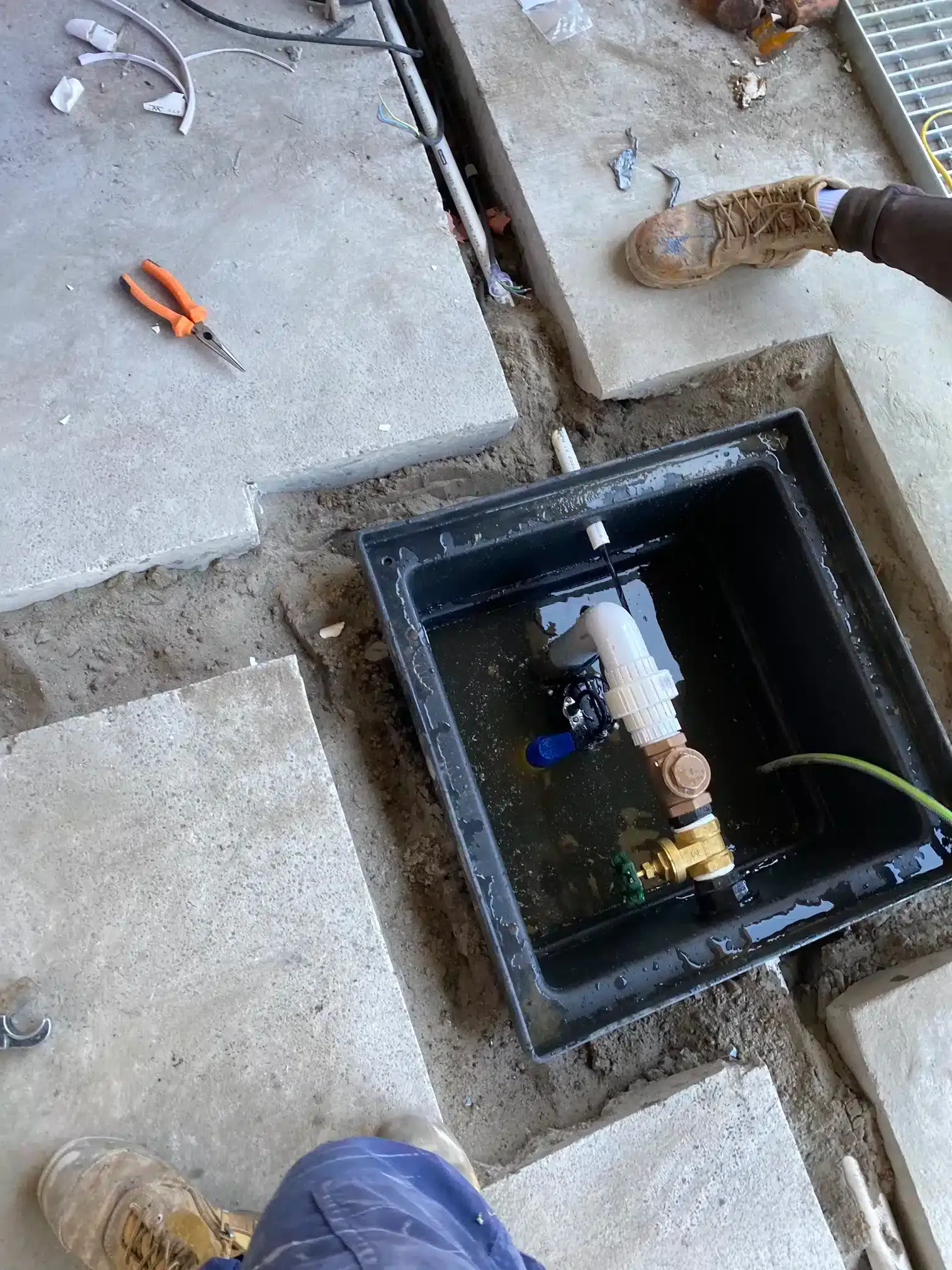 A black utility box embedded in a concrete ground, containing a plumbing valve assembly partially submerged in water.