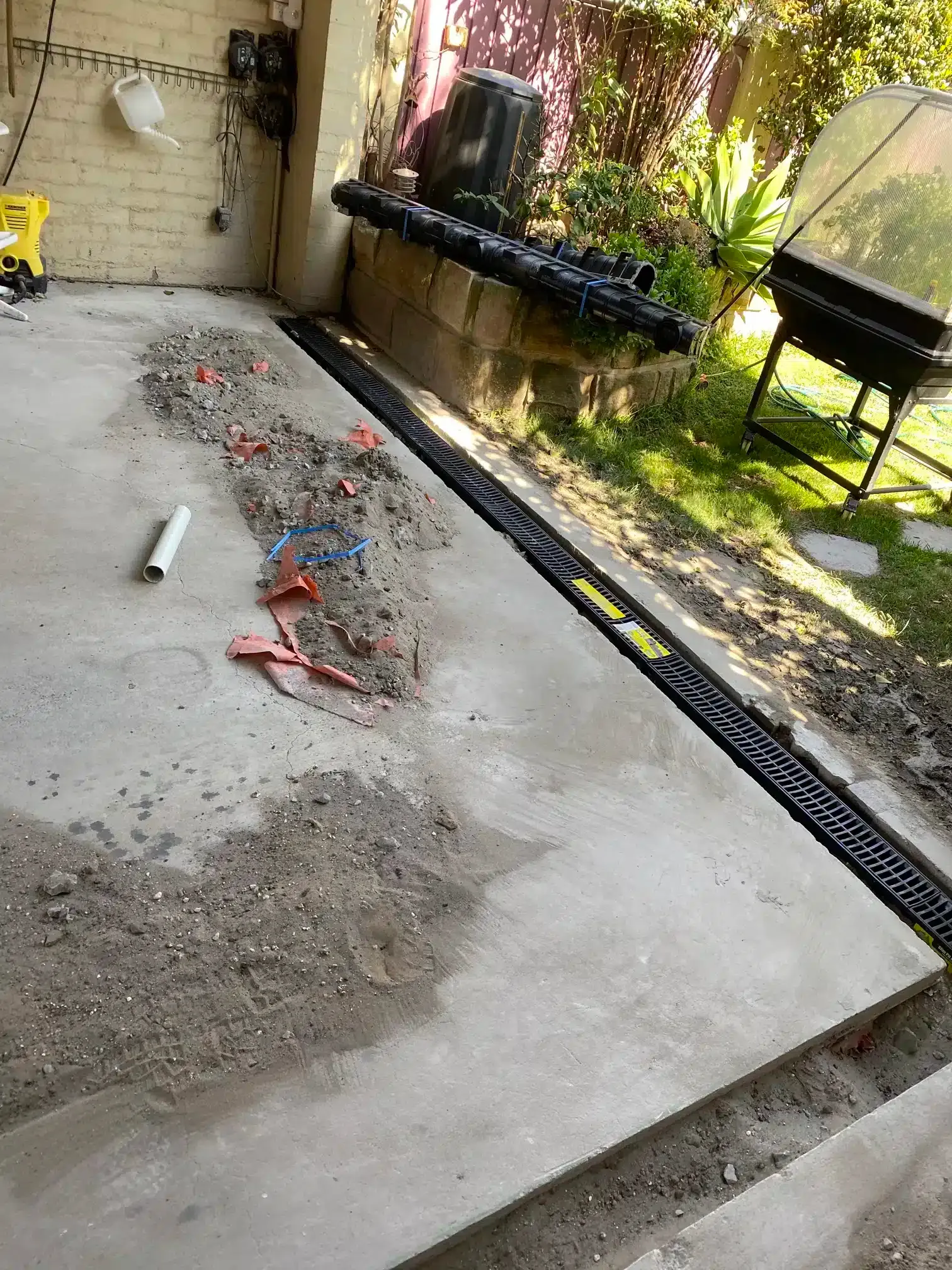 A black drainage channel installed in a concrete patio, with construction debris and a pipe nearby.