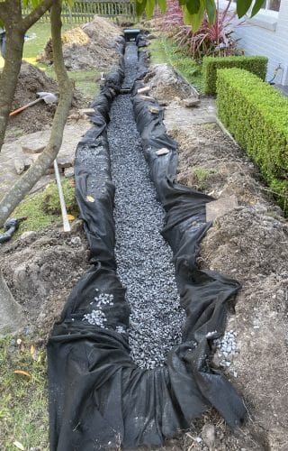 A trench lined with black landscape fabric and filled with dark gravel, situated in a residential yard for drainage.