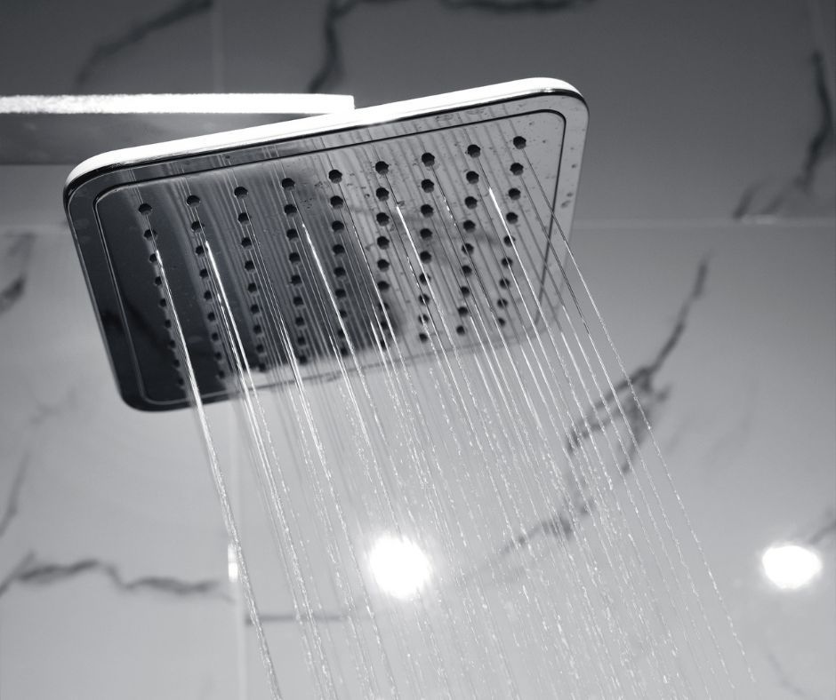 A rectangular shower head mounted on a marble-tiled wall, spraying multiple streams of water downward.