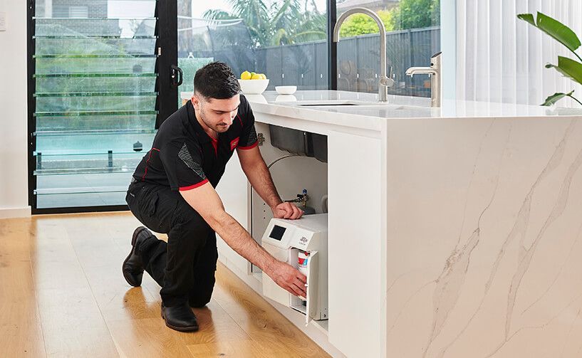 A technician kneels on a kitchen floor, installing or servicing a water filtration system unit under a marble countertop.