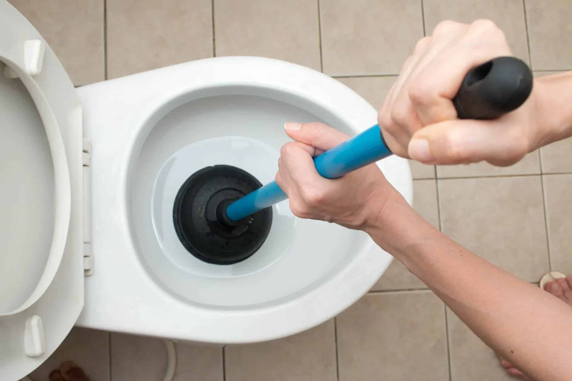 A person using a blue-handled plunger to unclog a white toilet in a tiled bathroom.