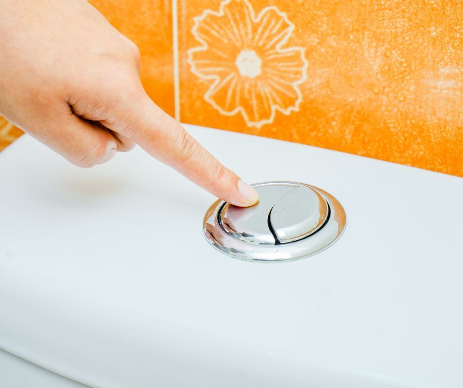 A finger pressing a split-button chrome toilet flush mechanism on a white tank against an orange tiled wall.