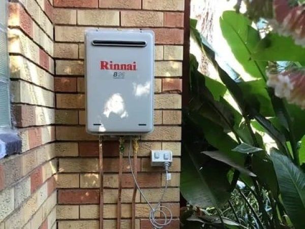 A Rinnai tankless gas water heater mounted on a brick exterior wall next to green foliage and an outdoor power outlet.