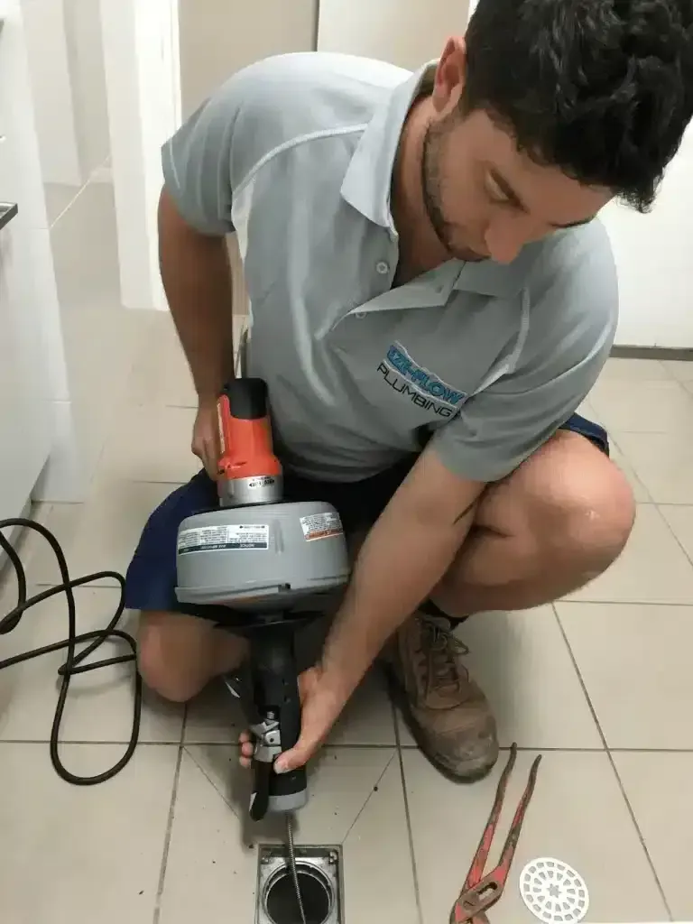 A technician kneeling on a tiled floor uses a powered plumbing snake to clear a drain, with tools visible nearby.