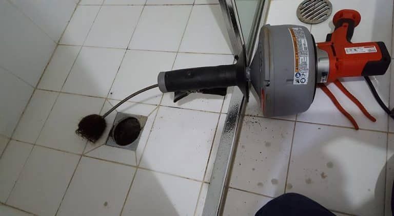 A red-and-gray motorized drain auger is positioned on a tiled floor, with its cable feeding into a shower floor drain.
