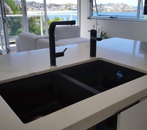 A black double-basin kitchen sink with two matching modern faucets, set in a white countertop overlooking a coastal view.