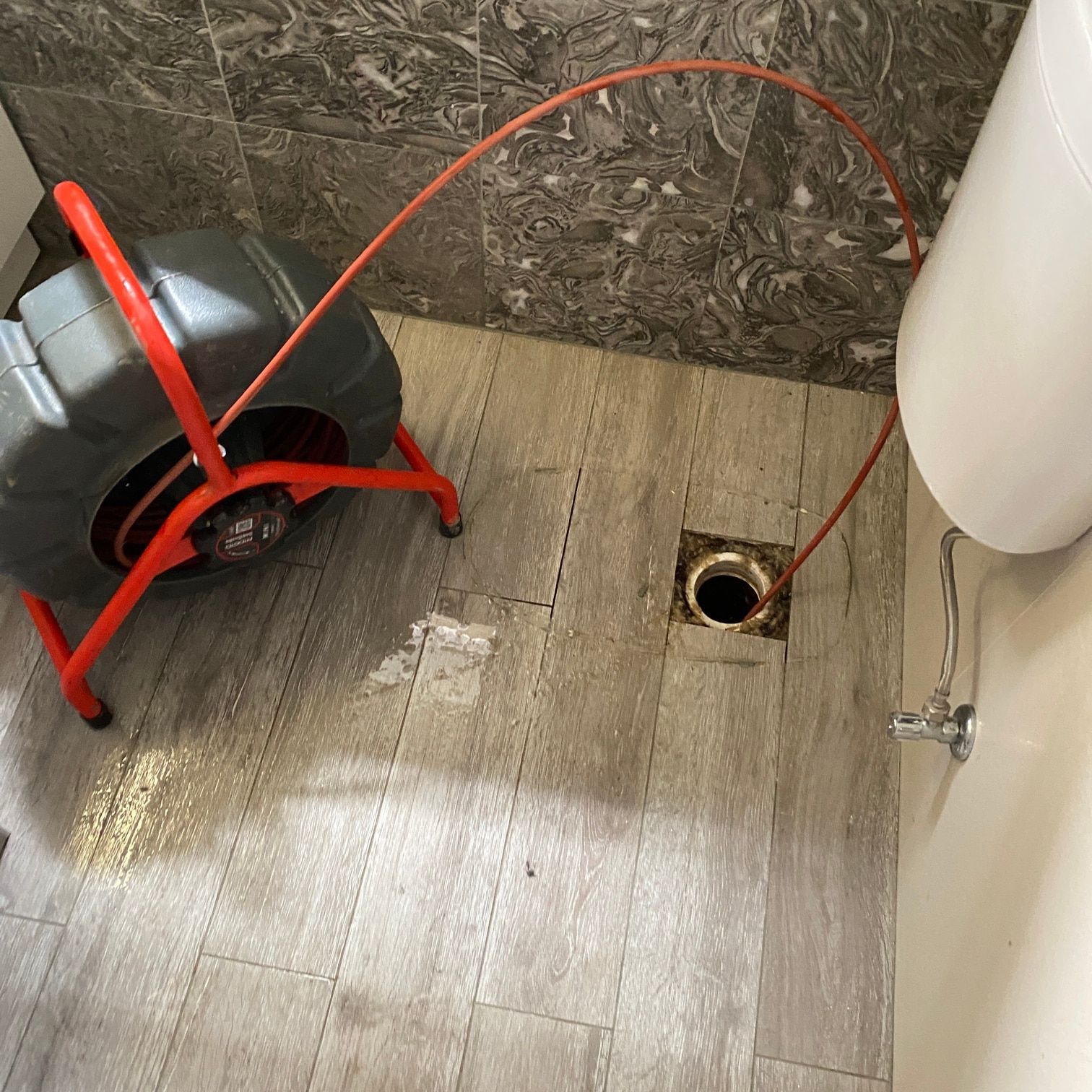 A red plumbing snake machine on a tiled floor with its orange cable fed into a floor drain near a toilet.