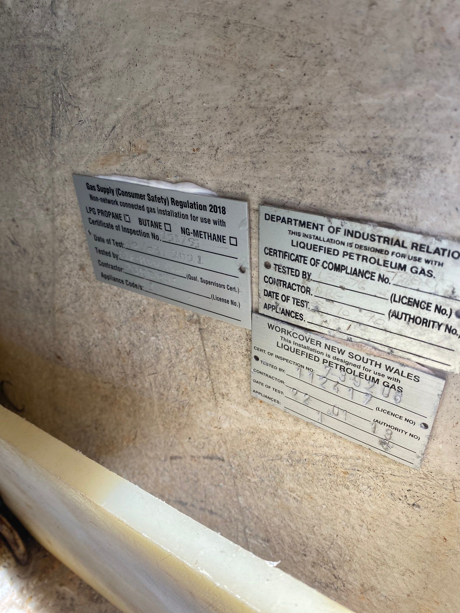 Three metal compliance plates for Liquefied Petroleum Gas mounted on a beige, textured wall.