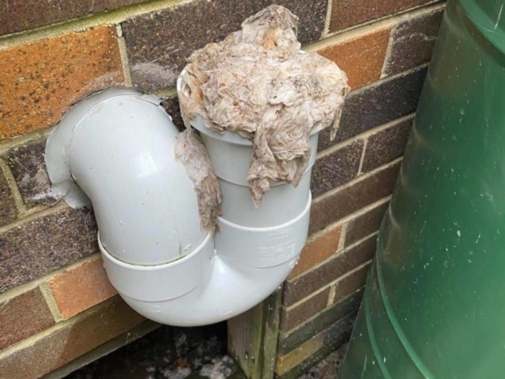 A white PVC P-trap pipe mounted on a brick wall, clogged and overflowing with a mass of fibrous, light-brown material.