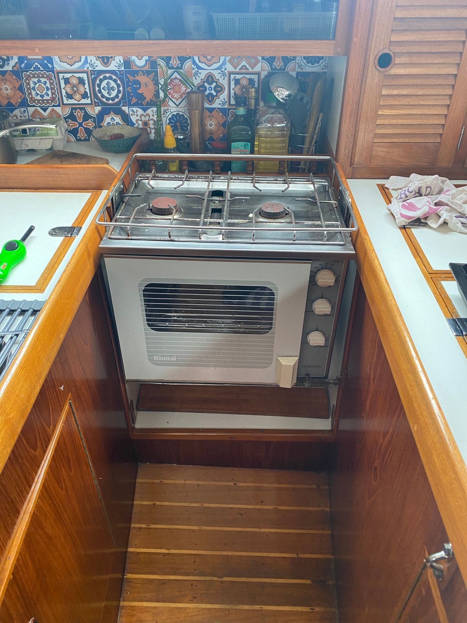 A stainless steel marine stove with two burners and an oven, set in a wood-paneled kitchen with a colorful tiled backsplash.