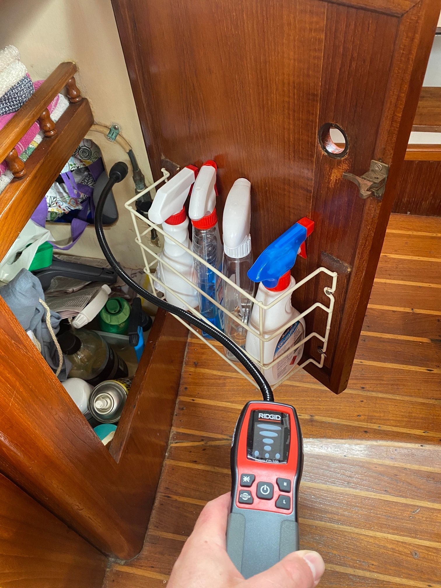 A handheld combustible gas detector with a probe held near cleaning spray bottles stored on the inside of a wood cabinet.