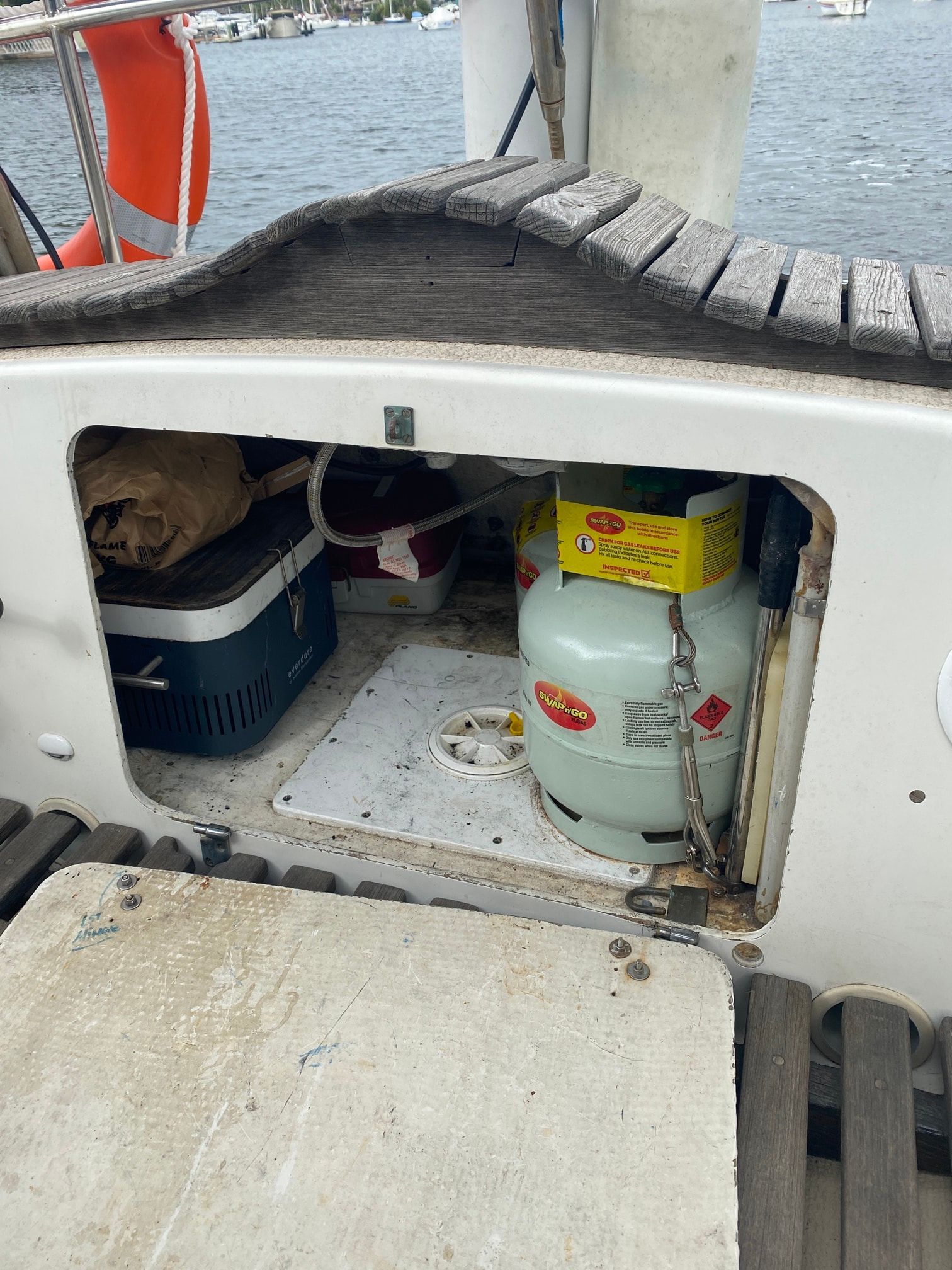 An open locker on a boat deck contains a propane tank, a small cooler, and various gear on a white floor.