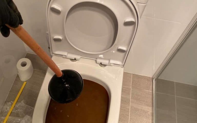 A gloved hand holds a black toilet plunger over a clogged, water-filled toilet bowl in a modern bathroom.
