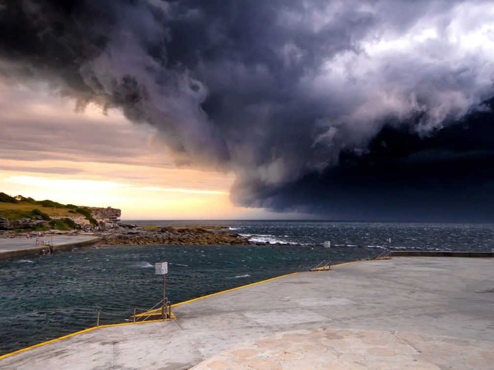 A dramatic storm front with dark, turbulent clouds looms over a calm sea, viewed from a concrete pier at dusk.