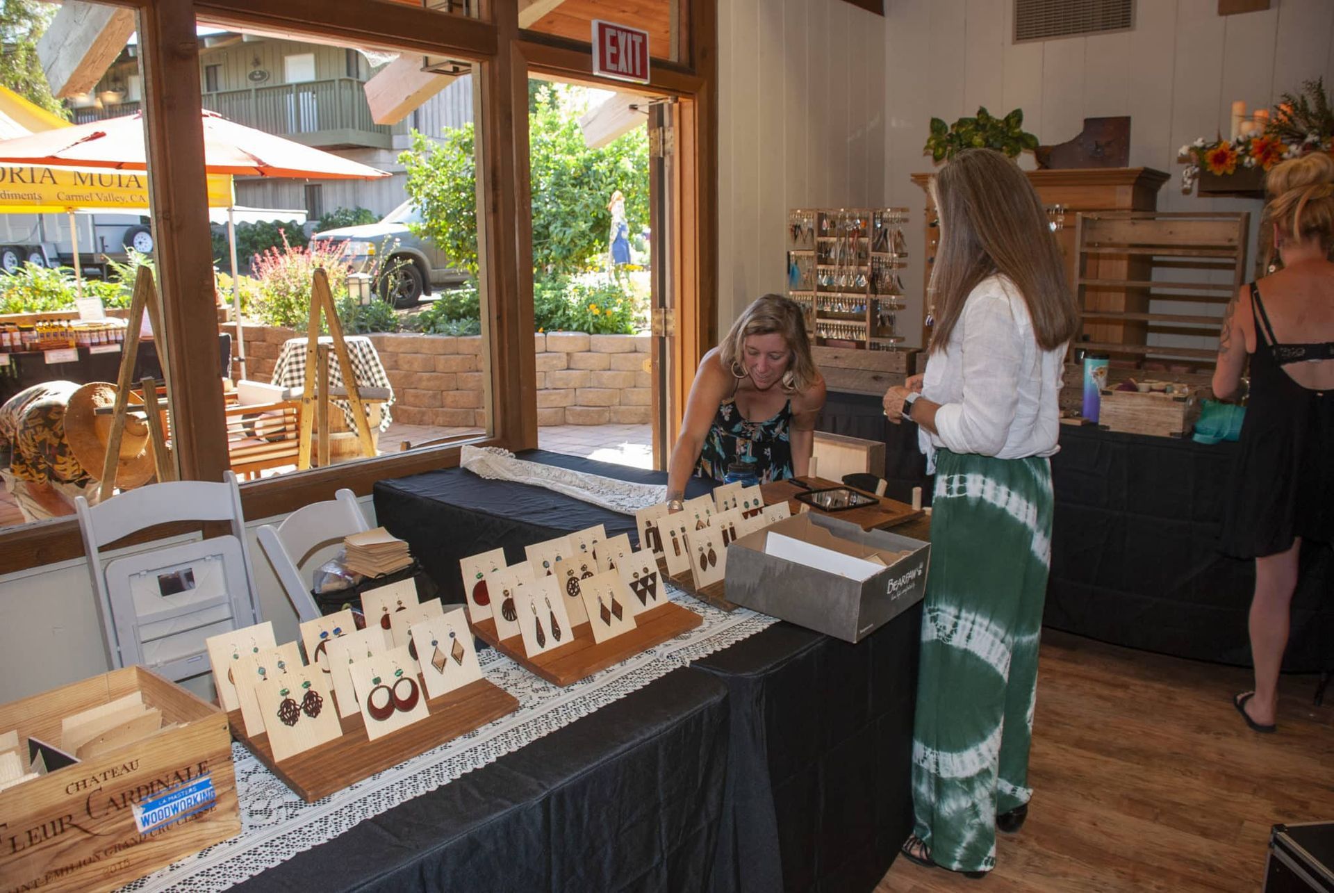 A table display with earrings on tan cardboard for sale in front of a large window and two women near the end of the table.
