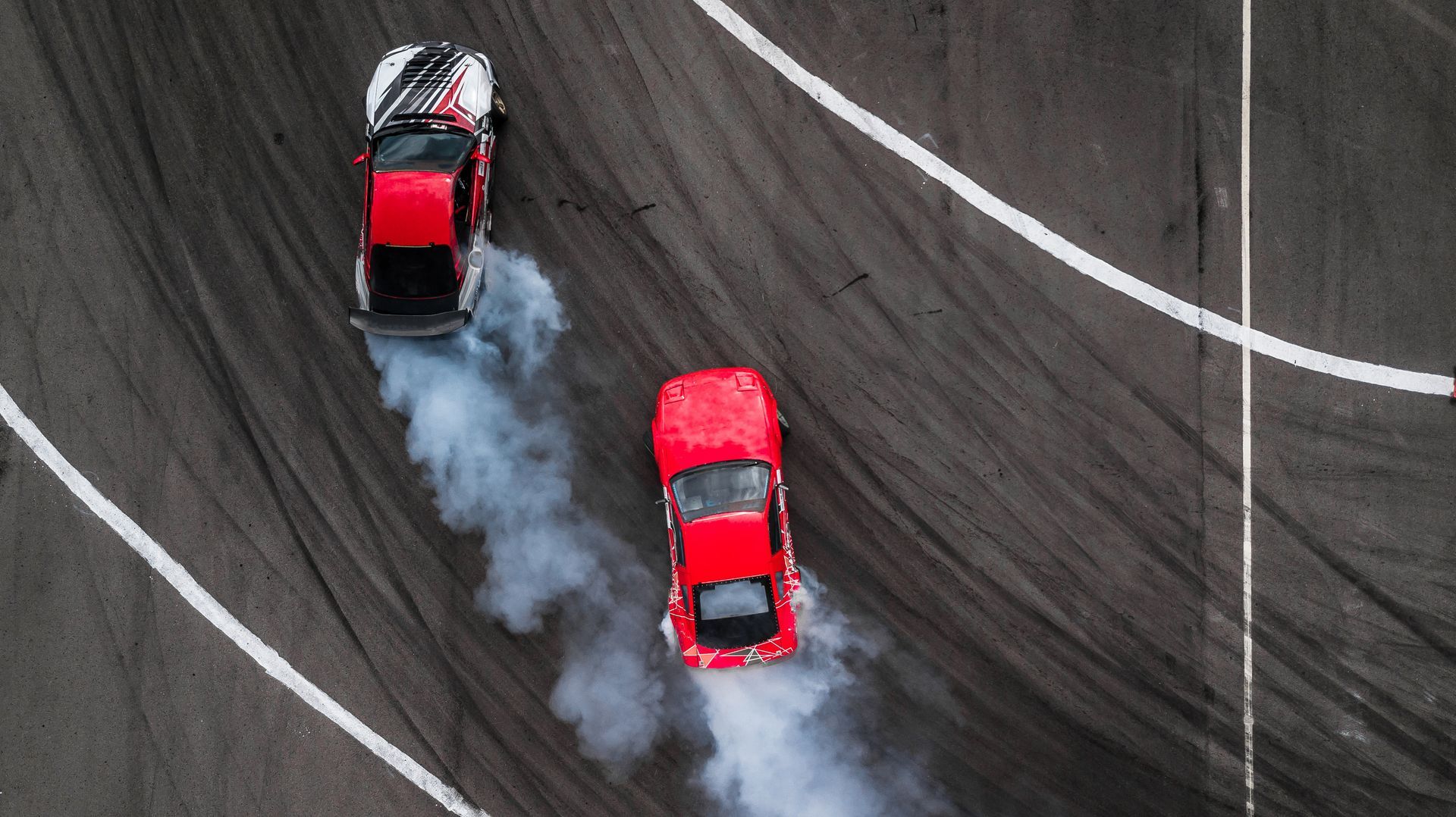 Two red cars drifting on a track, producing smoke.