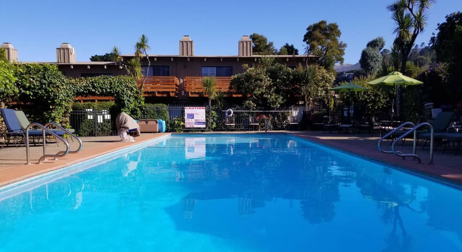 A large swimming pool is surrounded by chairs and umbrellas in front of a building.