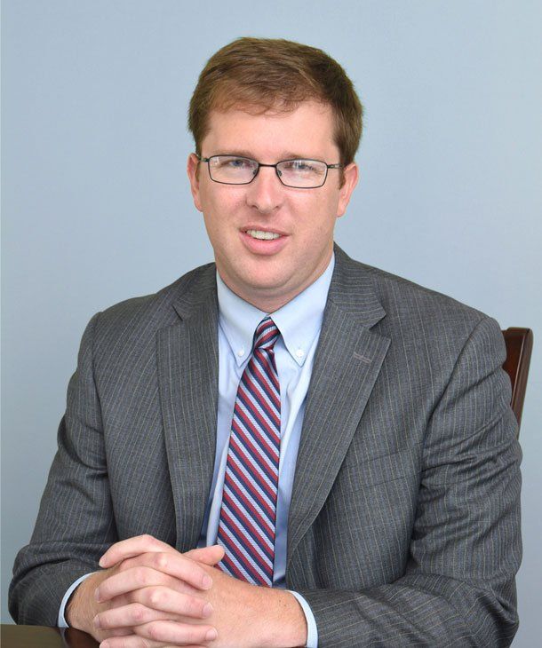 Man in a gray blazer and glasses, seated, hands clasped. Smiling, indoors.
