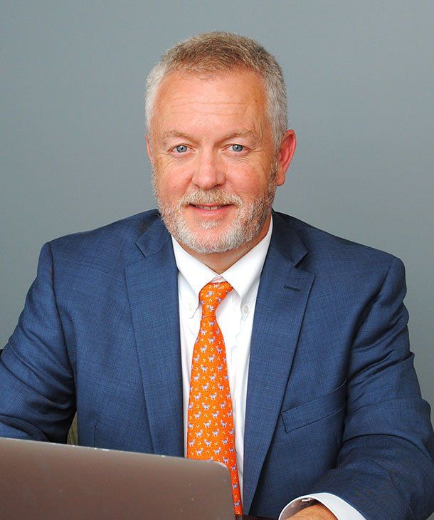 Man in blue suit, orange tie, smiling, seated at a desk with a laptop.